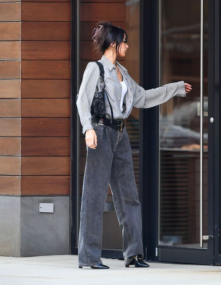 a woman in grey shirt and jeans walking down the street with her hand on her hip