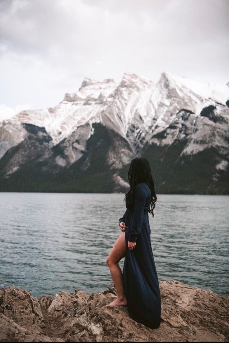 a woman sitting on top of a rock next to a body of water with snow covered mountains in the background