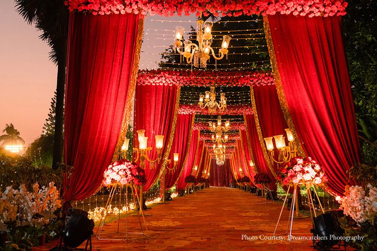 an outdoor wedding setup with red drapes and chandelier draped over the aisle