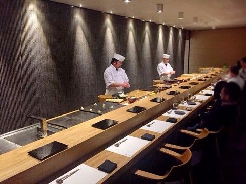 two chefs preparing food in a restaurant setting with wooden tables and chairs on the side