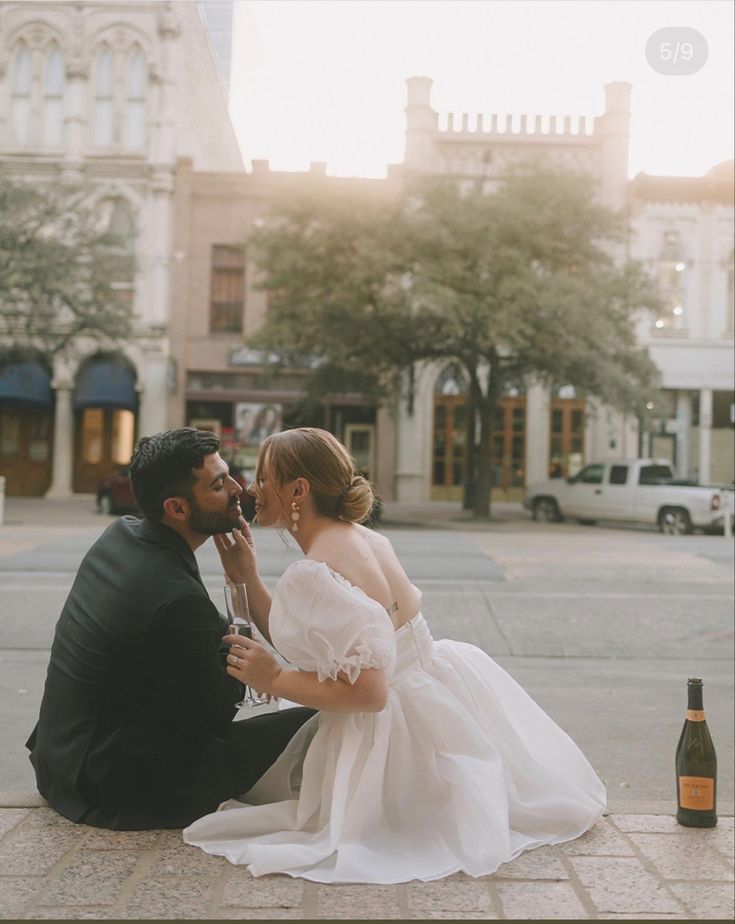 a man kneeling down next to a woman in a wedding dress on the ground with a bottle of wine