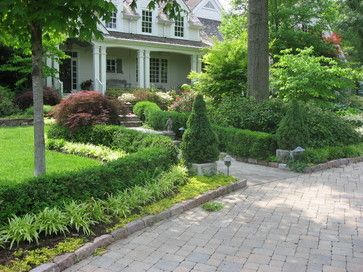 a house with lots of trees and bushes in front of it on a brick walkway