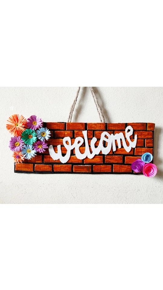 a sign that says welcome with flowers hanging from the side of a brick wall on a string