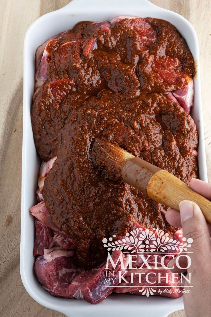 a person is spreading sauce on meat in a casserole dish with a wooden spatula