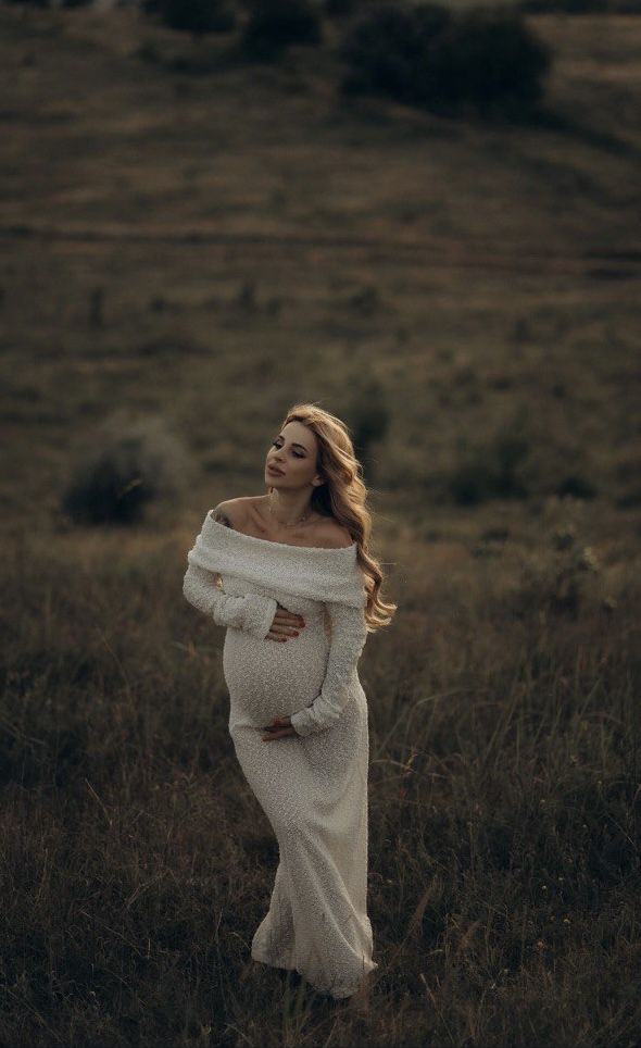 a pregnant woman standing in a field with her arms wrapped around her waist and looking off to the side