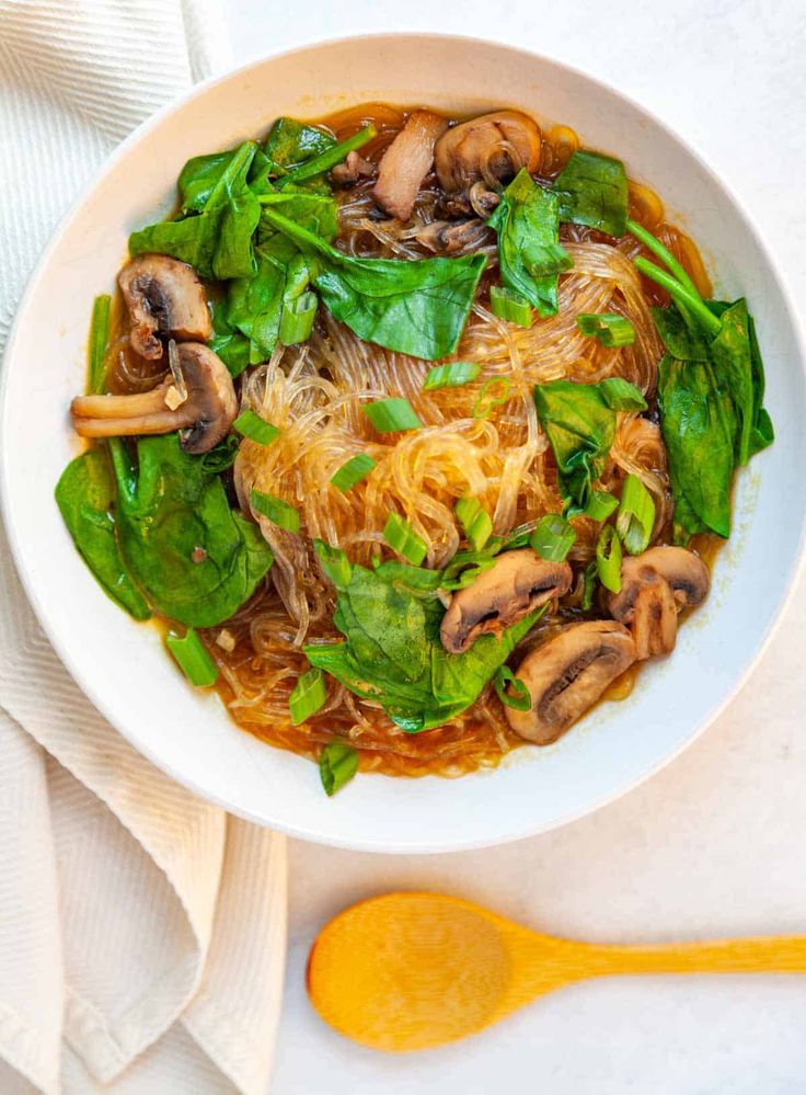 a white bowl filled with noodles, mushrooms and spinach on top of a table