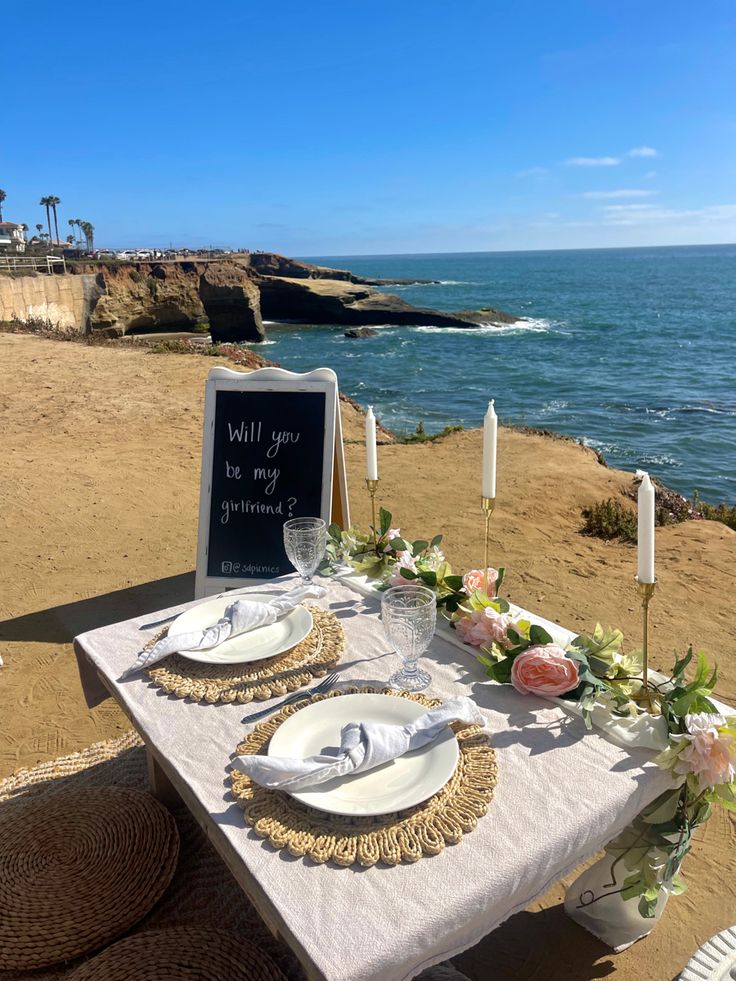 a table set up on the beach with flowers and candles for an outdoor dinner party