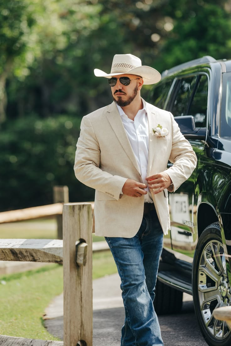 a man in a cowboy hat and sunglasses standing next to a black truck wearing a tan blazer