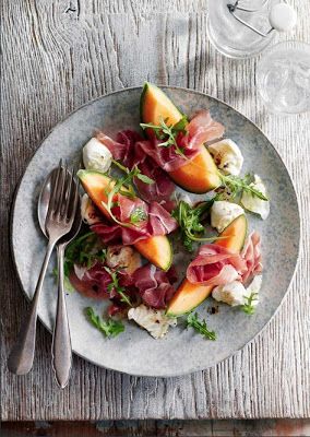 a white plate topped with fruit and meat on top of a wooden table next to silverware