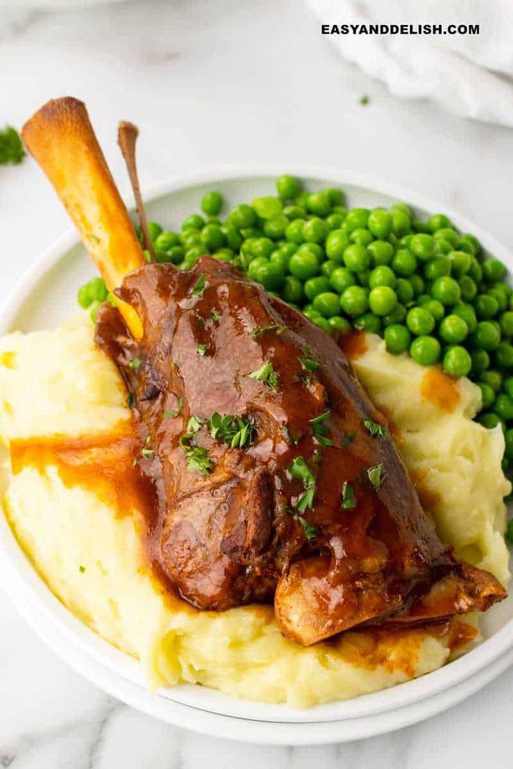 a white plate topped with meat and mashed potatoes next to green peas on a marble table