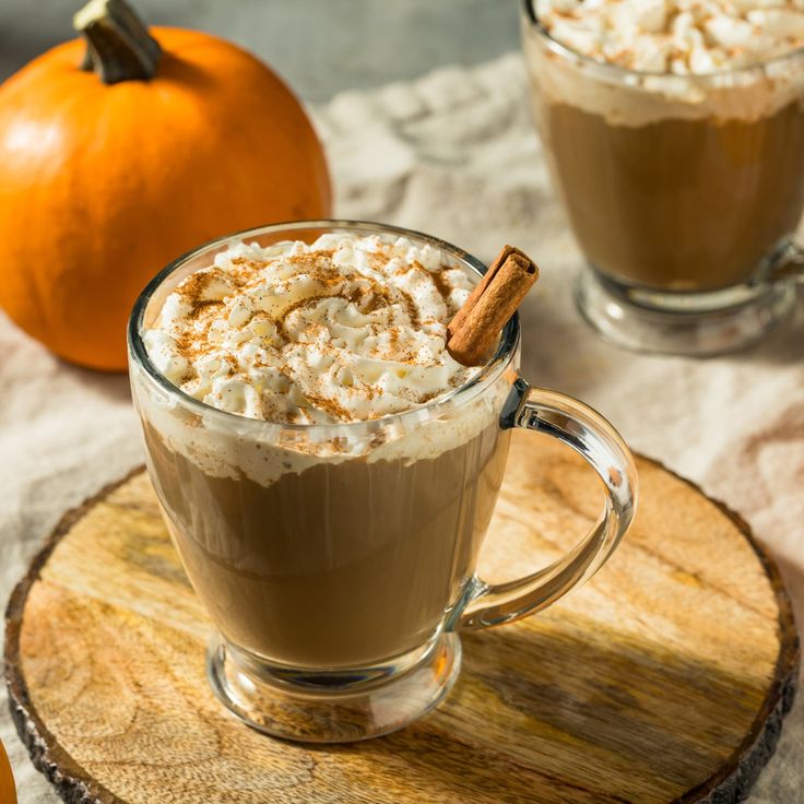 two mugs of hot chocolate with whipped cream and cinnamon on a wooden tray next to an orange pumpkin