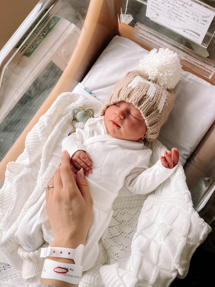 a newborn baby in a hospital bed being held by a person's hand and wearing a knitted hat