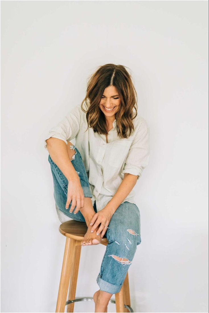 a woman sitting on top of a wooden stool next to a white wall and smiling