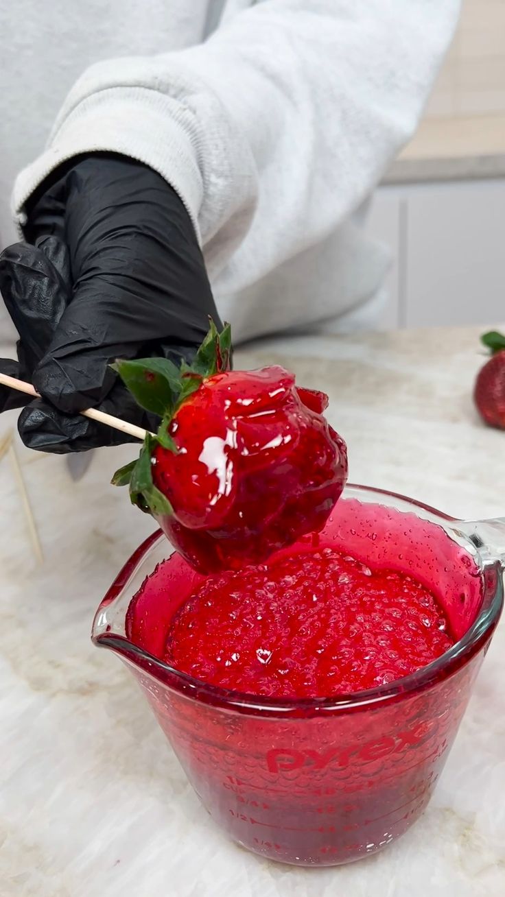 a person in black gloves holding a strawberry over a red liquid filled glass bowl with strawberries on the side