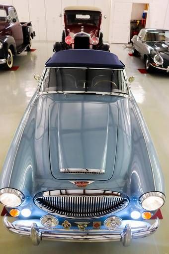 an old blue car is parked in a garage with other classic cars behind it and on the floor