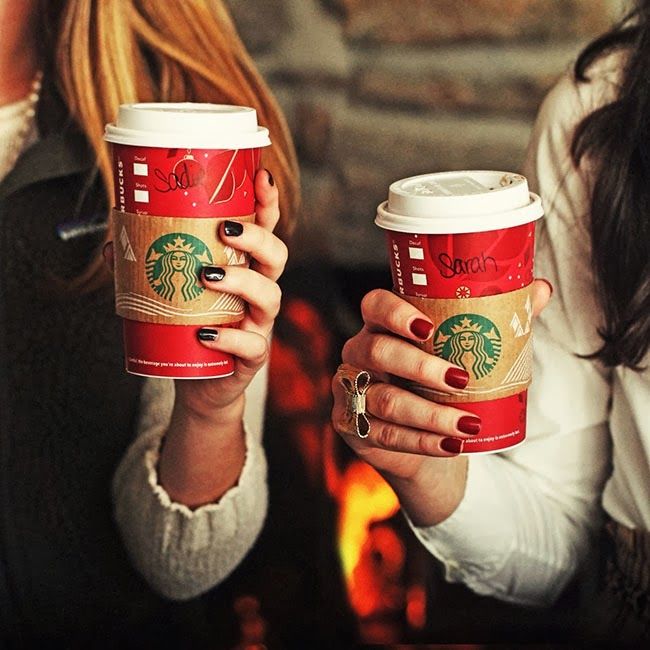 two women holding up starbucks cups in front of a fire place