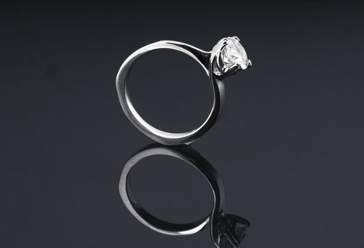 an engagement ring with a single diamond in the center on a reflective surface, against a black background