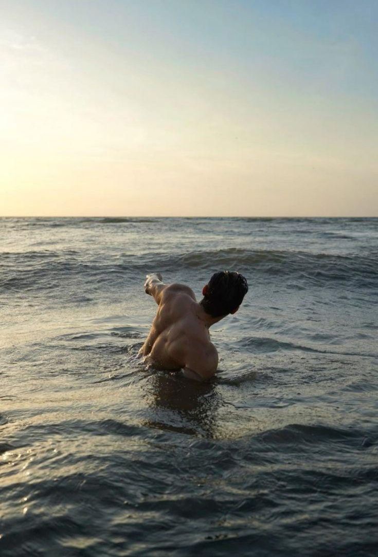 a shirtless man swimming in the ocean with his back to the camera and arms outstretched