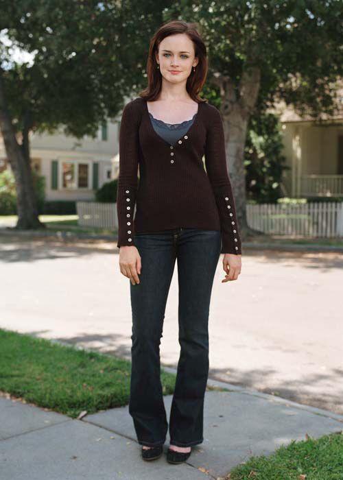 a woman is standing on the sidewalk talking on her cell phone while wearing jeans and a sweater