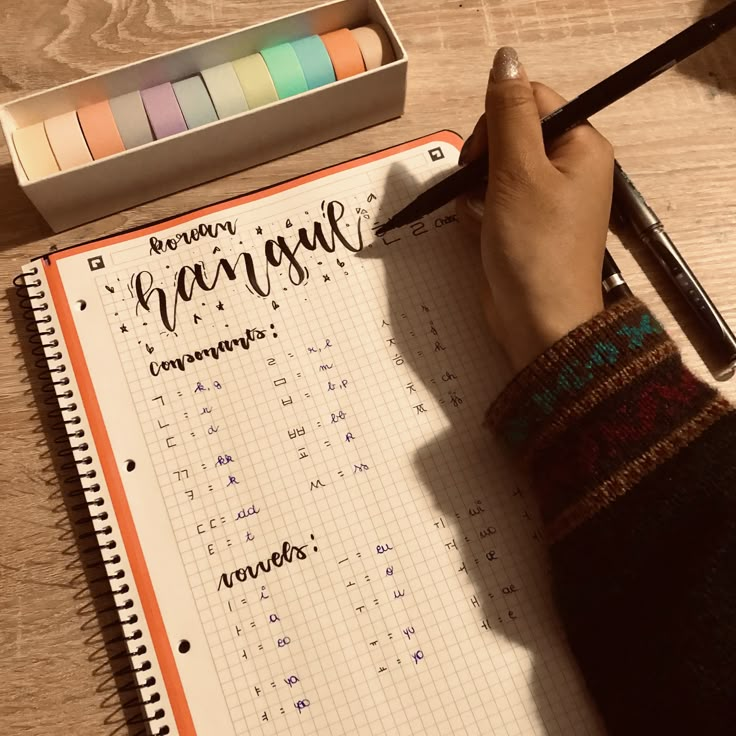 a person writing on a notebook with markers and pencils next to it, while holding a pen in their left hand