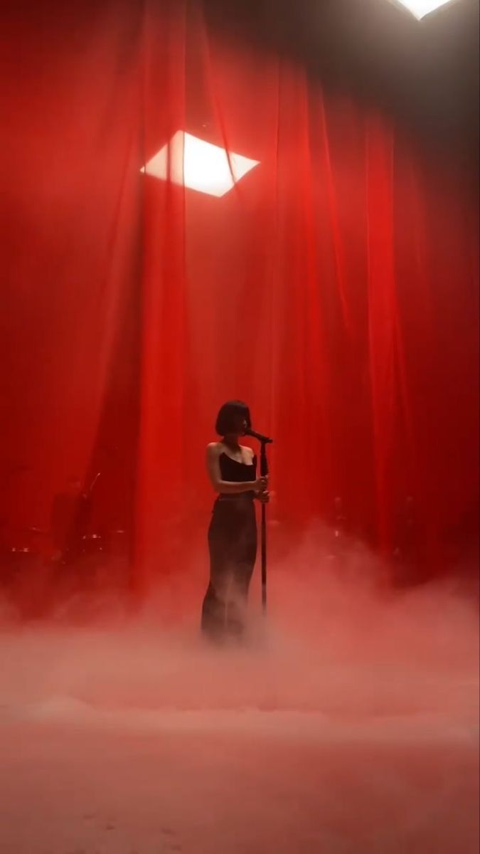 a woman standing in front of a red curtain on stage with a microphone behind her