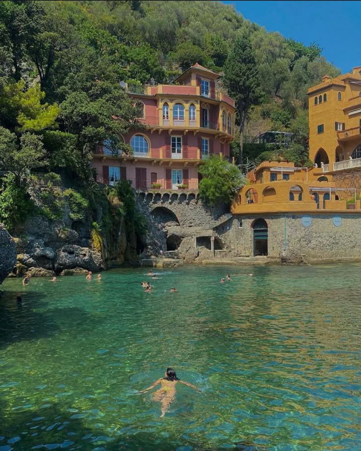 people are swimming in the clear blue water near some yellow buildings and green trees on top of a hill