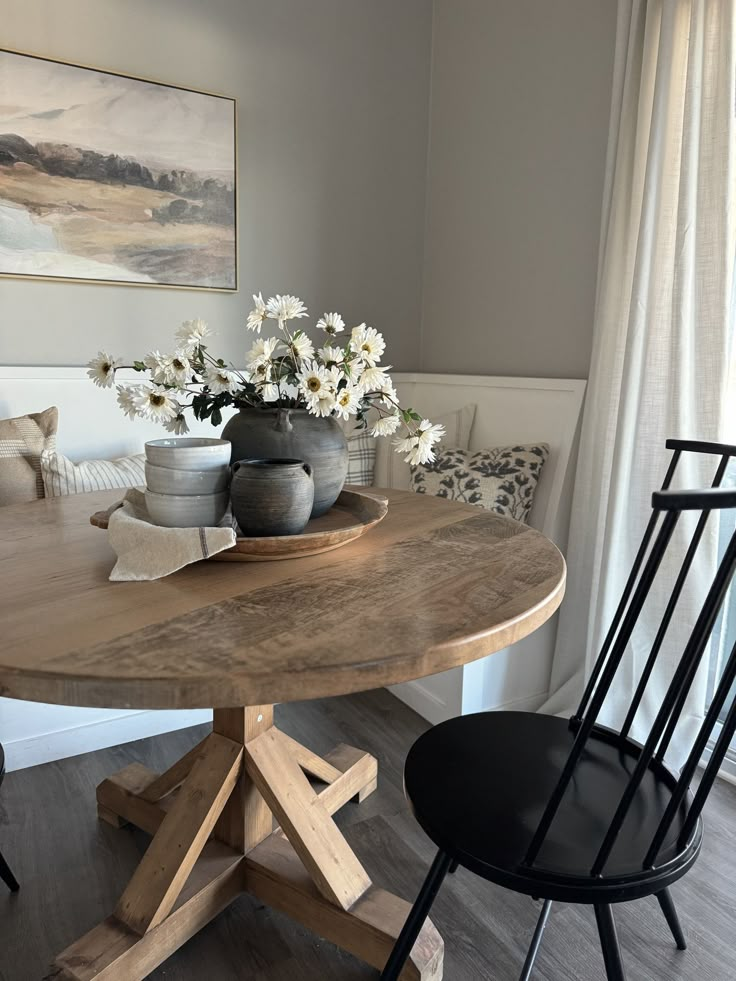 a wooden table sitting in front of a window next to a chair and vase with flowers on it