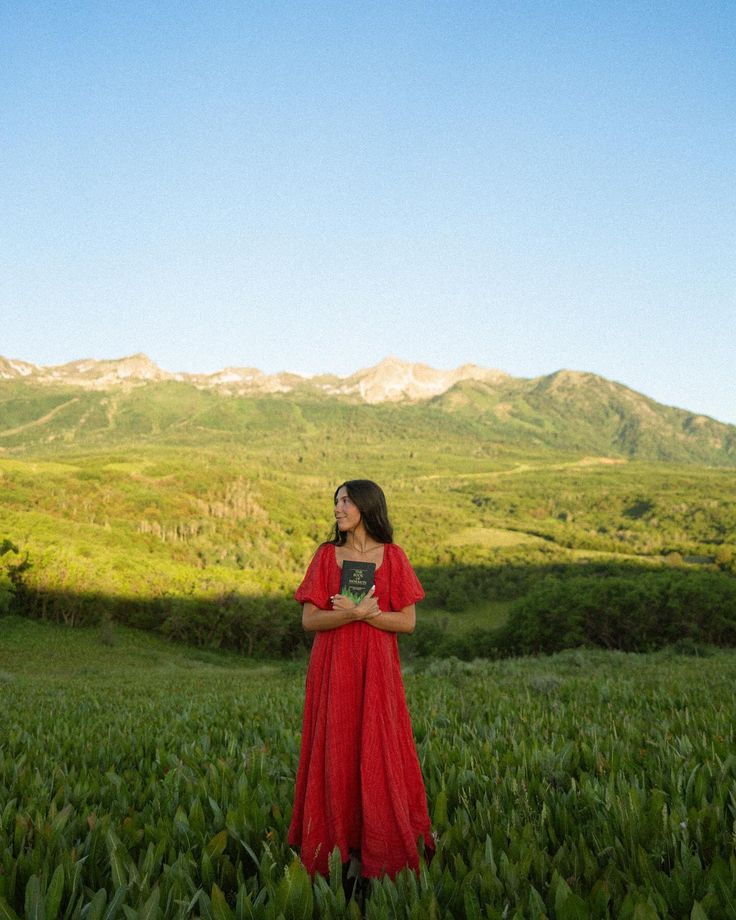 a woman in a red dress standing in the middle of a field with mountains behind her