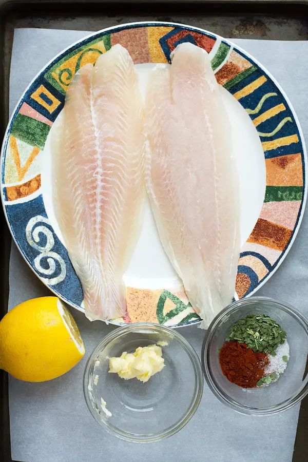 two fish fillets on a plate with spices and seasoning next to lemons