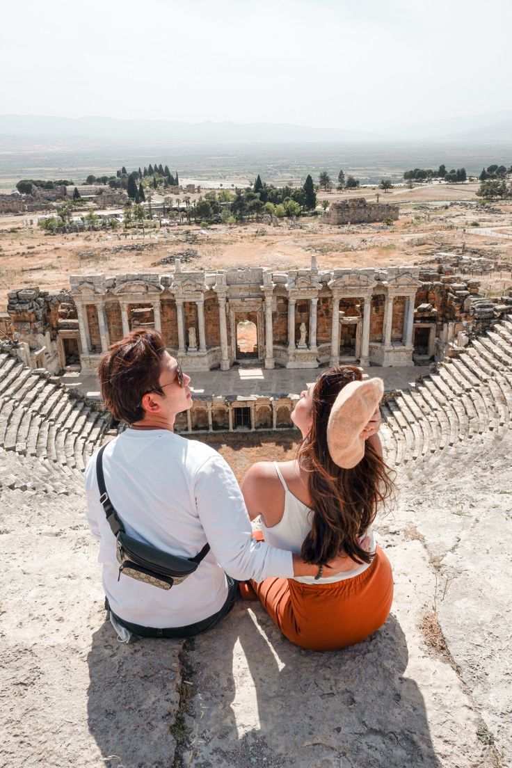 a man and woman sitting on the ground looking at an ancient amphite with ruins in the background