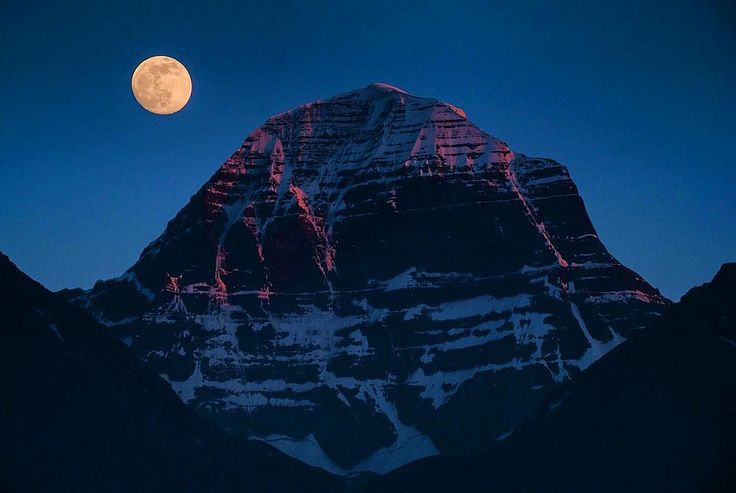 the full moon is setting behind a snowy mountain