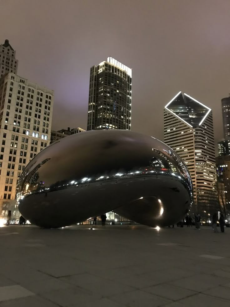 a large metal object in the middle of a city at night with skyscrapers behind it