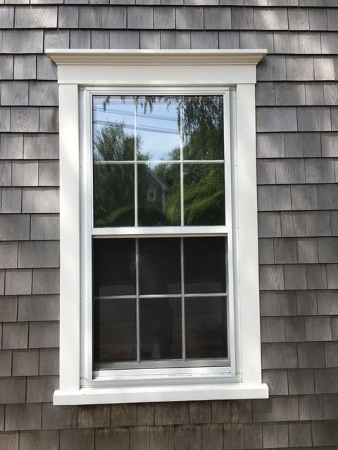 an open window on the side of a gray shingled house with trees in the background