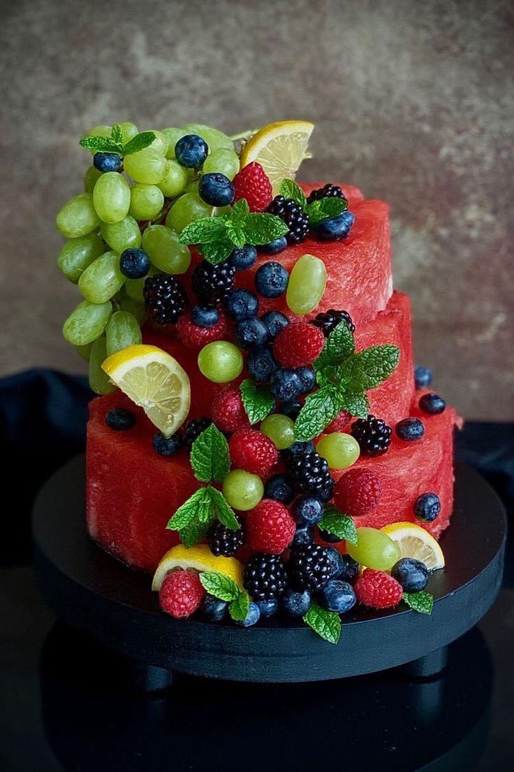 a watermelon cake with berries, grapes and limes on top sits on a black plate