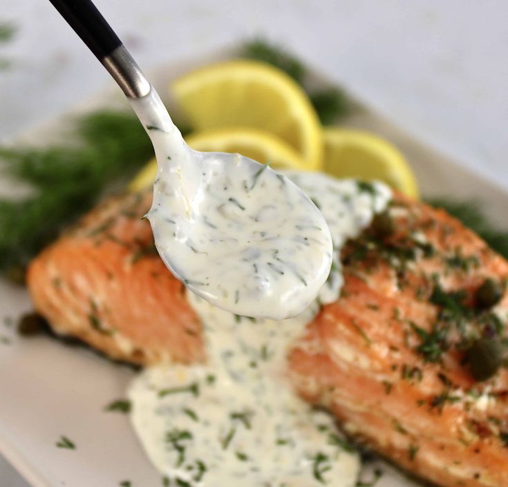 a spoon full of cream sauce being held over salmon on a plate with lemon wedges and parsley