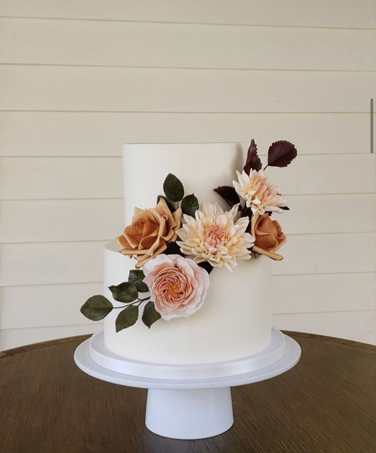 there is a white cake with flowers on the top and leaves on the bottom, sitting on a wooden table