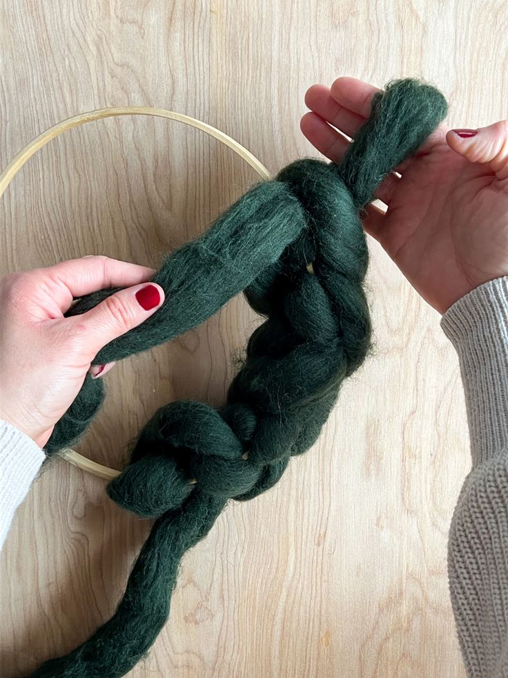 two hands holding a piece of green yarn in front of a hoop on a wooden surface