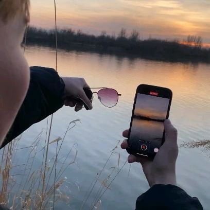 a person holding up a cell phone next to a body of water with a sunset in the background