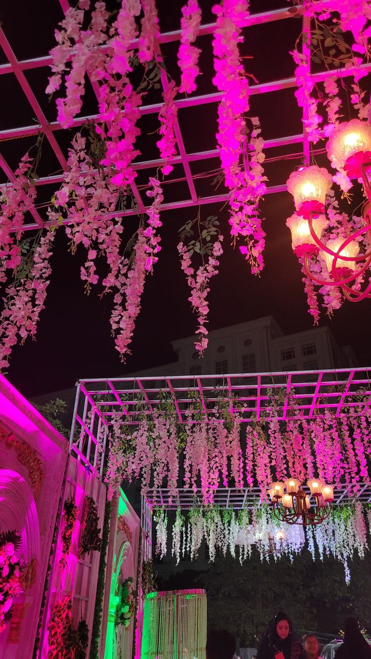 pink flowers are hanging from the ceiling in an outdoor venue at night with lights and chandeliers