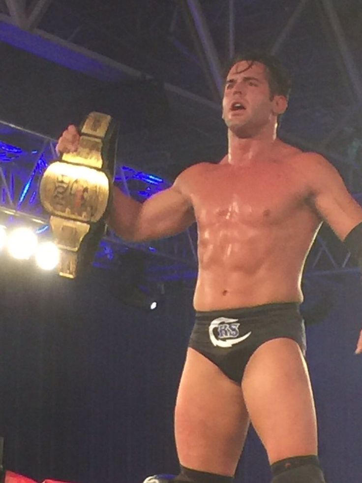 a shirtless man standing on top of a wrestling ring holding a gold belt in his right hand