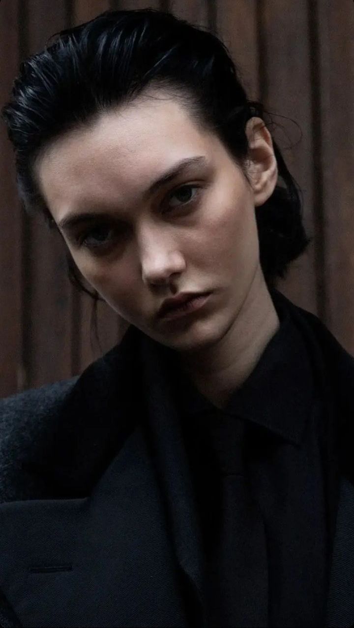 a woman with black hair is wearing a suit and looking off to the side while standing in front of a wooden wall