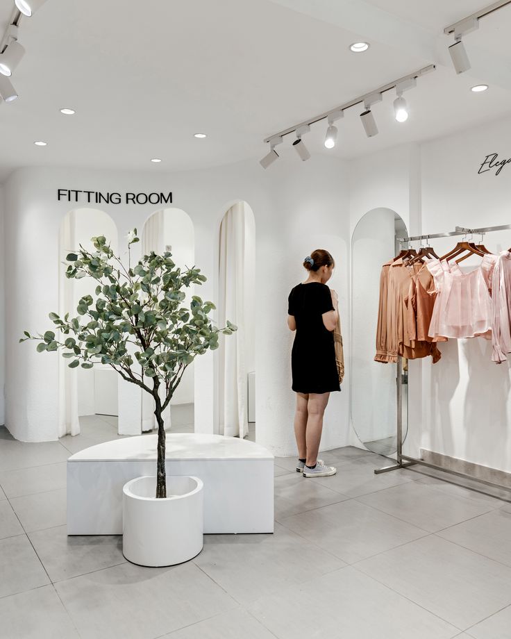 a woman standing in front of a clothing rack with clothes on hangers and a potted tree