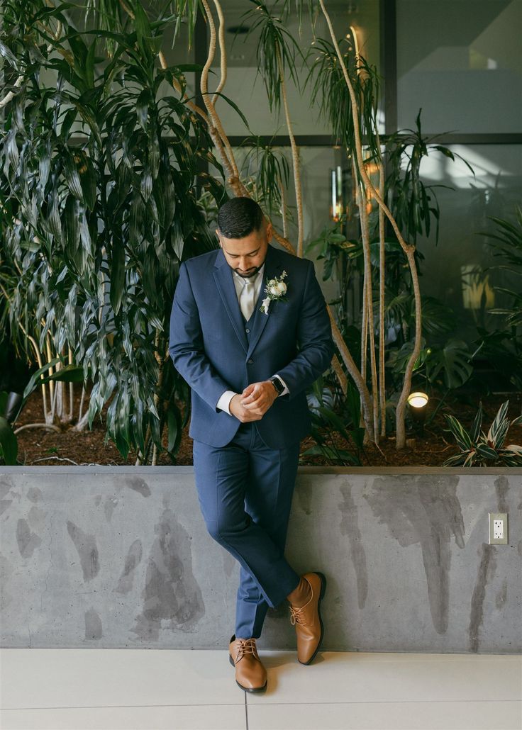 a man in a blue suit leaning against a wall looking down at his cell phone