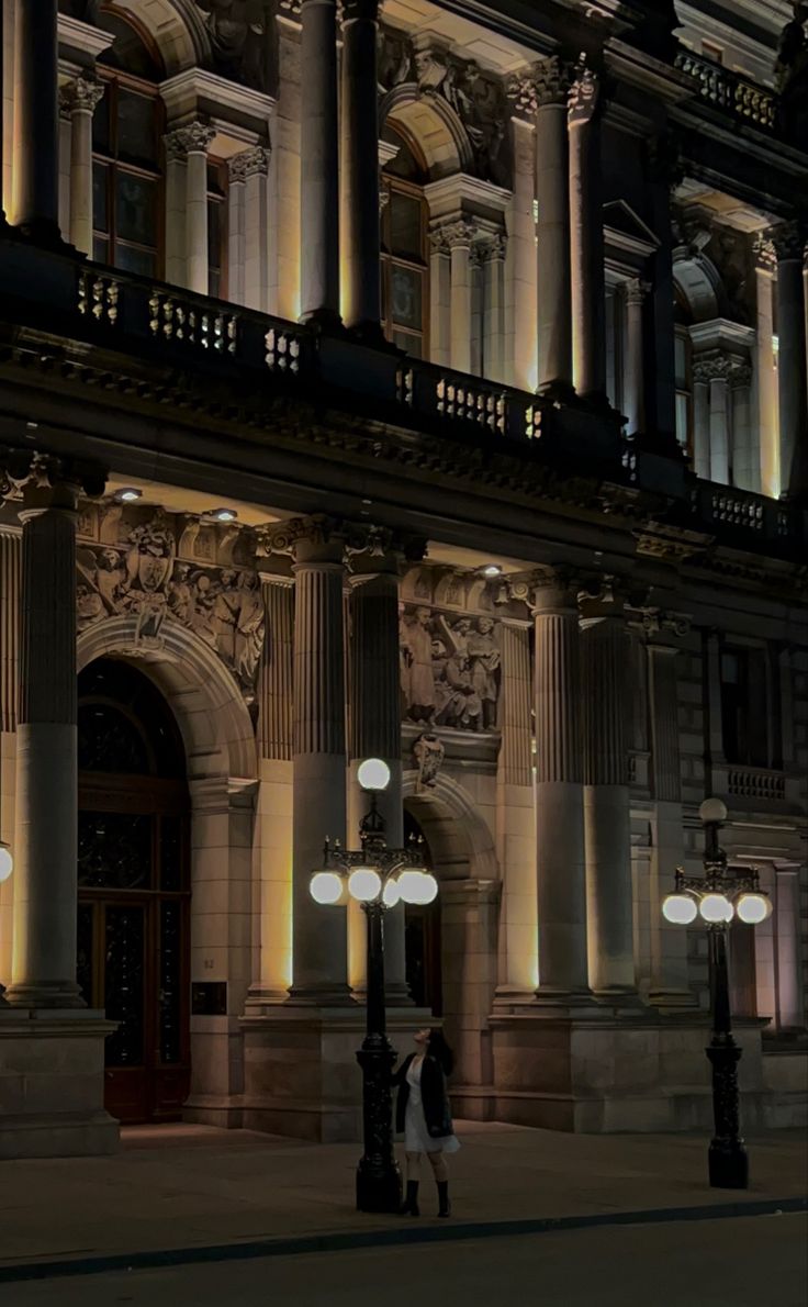 an old building lit up at night with street lamps in front of it and people walking by