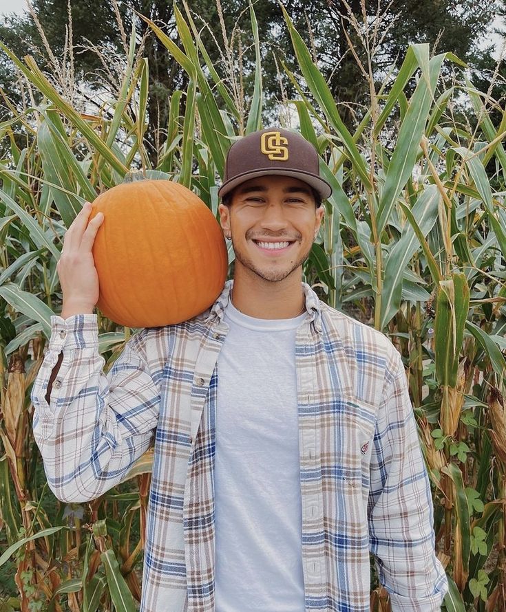 a man holding a pumpkin in front of a corn field with trees and bushes behind him