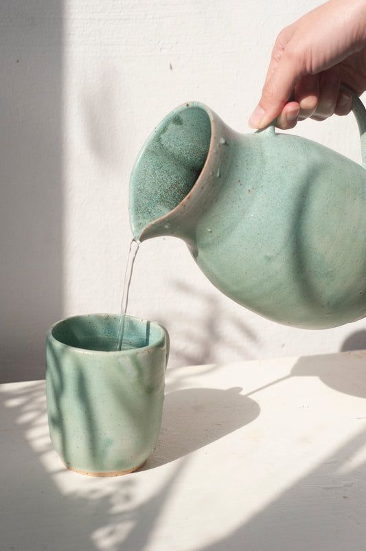 a person pours water into a cup from a teapot on a table in front of a white wall