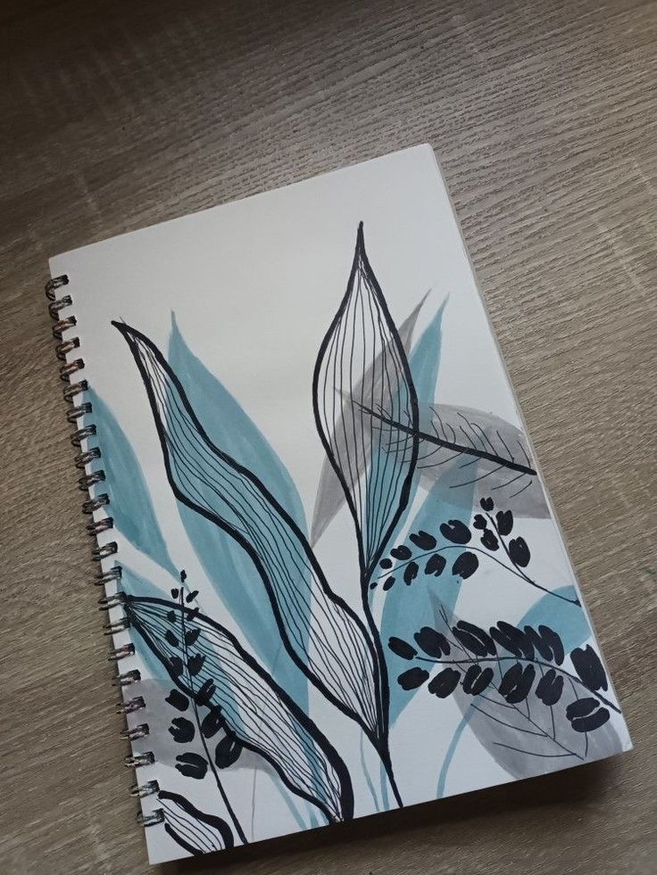 a spiral notebook with blue leaves on it sitting on top of a wooden table next to a pen