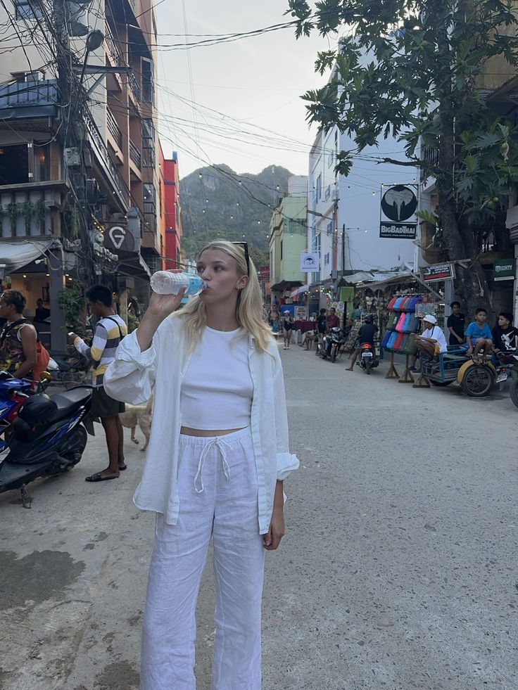 a woman standing in the middle of a street drinking from a bottle while wearing white pants