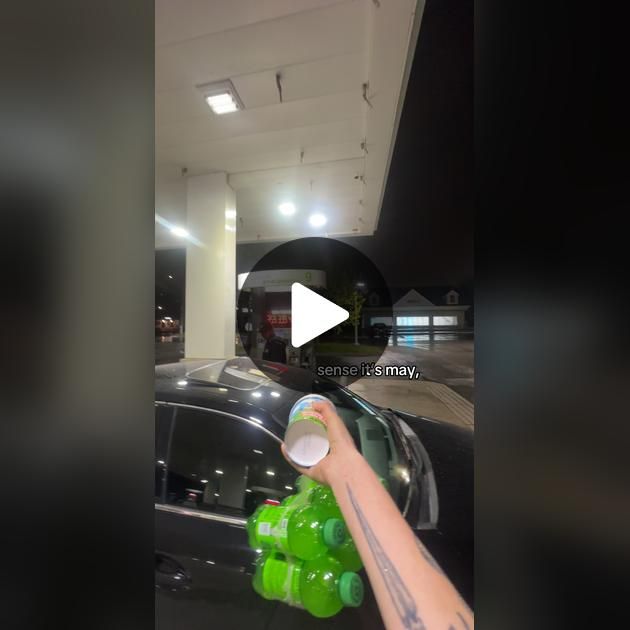 a hand is holding a green plastic bottle in front of a black car at a gas station