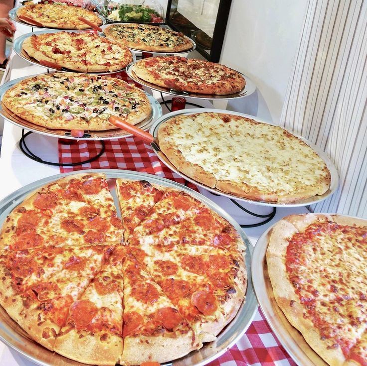 many different pizzas on pans lined up on a table with red and white checkered cloth